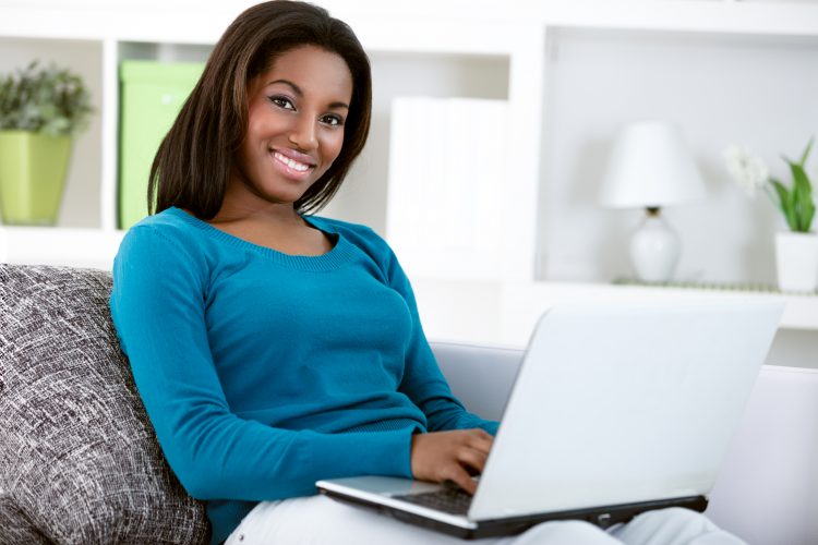 young black girl with laptop computer, sitting in bright living room, looking at camera.