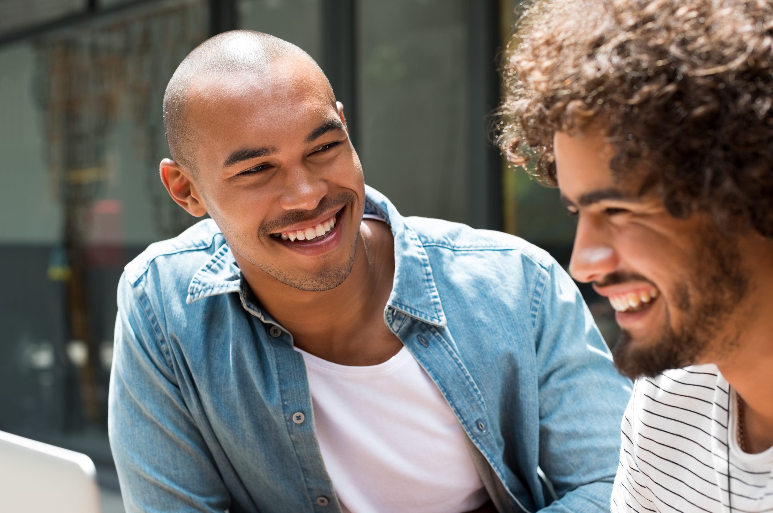 Close up face of best friends sitting and laughing. Happy african man smiling and looking at beard friend with curly hair. College students enjoying and relax outdoor.